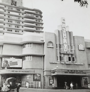 A black and white photograph of an old cinema building labeled 'Cathay' with art deco architectural features. The marquee advertises a movie titled 'The Ghost Breakers'. Pedestrians walk by on the street in front of the cinema.