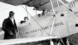Three individuals dressed in early 20th-century aviator attire are seen with a vintage biplane labeled 'Mildura Flying School'. Two people are seated in the cockpit, while the third stands beside the plane. The image suggests a historical aviation context.