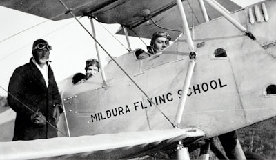 Three individuals dressed in early 20th-century aviator attire are seen with a vintage biplane labeled 'Mildura Flying School'. Two people are seated in the cockpit, while the third stands beside the plane. The image suggests a historical aviation context.