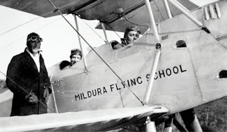 Three individuals dressed in early 20th-century aviator attire are seen with a vintage biplane labeled 'Mildura Flying School'. Two people are seated in the cockpit, while the third stands beside the plane. The image suggests a historical aviation context.