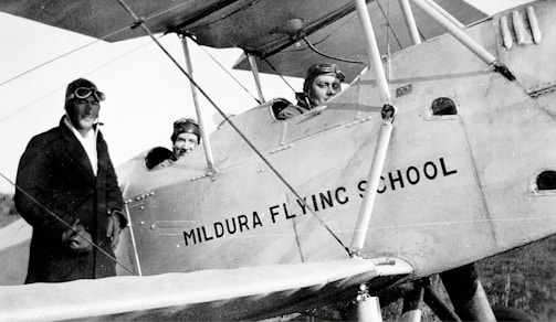 Three individuals dressed in early 20th-century aviator attire are seen with a vintage biplane labeled 'Mildura Flying School'. Two people are seated in the cockpit, while the third stands beside the plane. The image suggests a historical aviation context.