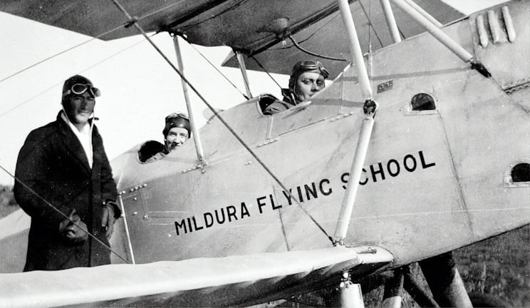 Three individuals dressed in early 20th-century aviator attire are seen with a vintage biplane labeled 'Mildura Flying School'. Two people are seated in the cockpit, while the third stands beside the plane. The image suggests a historical aviation context.