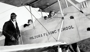 Three individuals dressed in early 20th-century aviator attire are seen with a vintage biplane labeled 'Mildura Flying School'. Two people are seated in the cockpit, while the third stands beside the plane. The image suggests a historical aviation context.