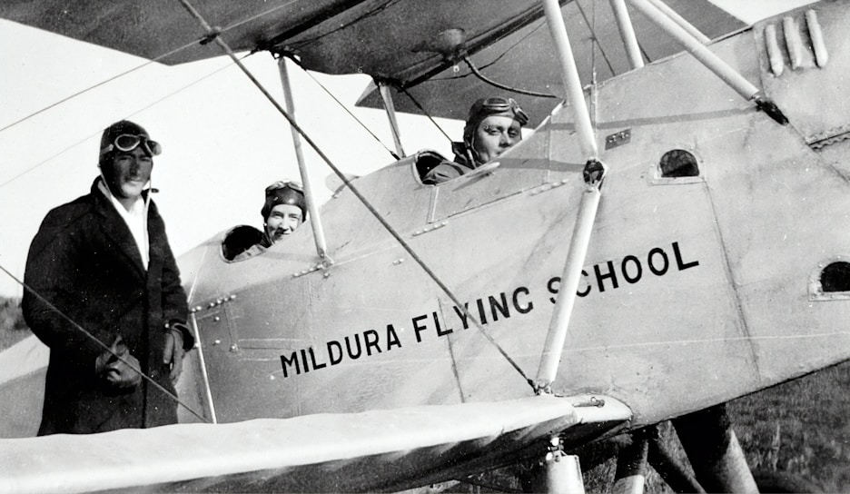 Three individuals dressed in early 20th-century aviator attire are seen with a vintage biplane labeled 'Mildura Flying School'. Two people are seated in the cockpit, while the third stands beside the plane. The image suggests a historical aviation context.