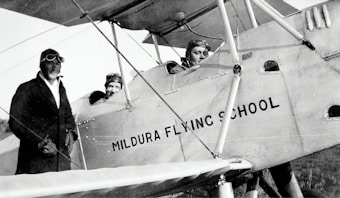 Three individuals dressed in early 20th-century aviator attire are seen with a vintage biplane labeled 'Mildura Flying School'. Two people are seated in the cockpit, while the third stands beside the plane. The image suggests a historical aviation context.