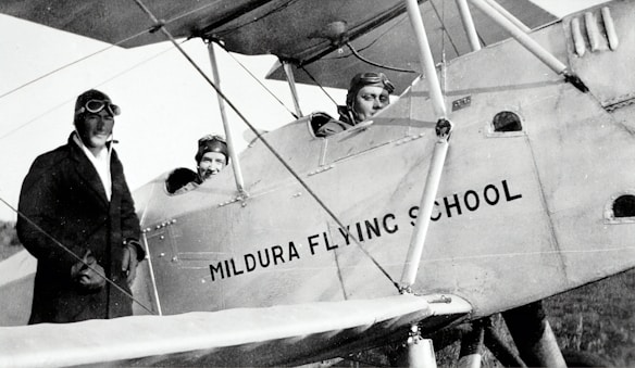 Three individuals dressed in early 20th-century aviator attire are seen with a vintage biplane labeled 'Mildura Flying School'. Two people are seated in the cockpit, while the third stands beside the plane. The image suggests a historical aviation context.