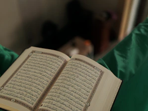 Close-up of hands holding an open Quran with elegant gold and green design elements softly blurred behind.