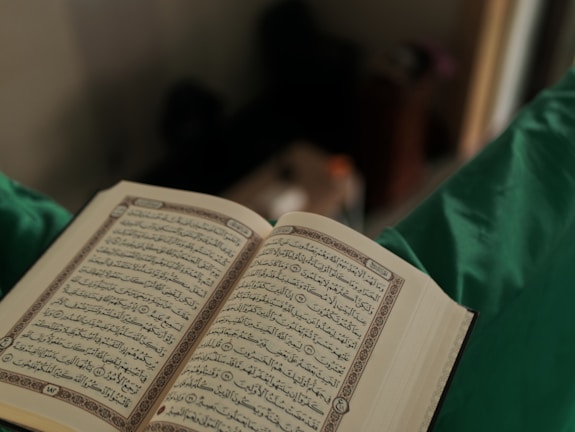 An open book with Arabic script is held in a person's hands, set against a blurred background. The page layout and text density suggest a religious or holy book.