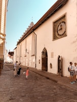 A historical street scene features a group of people, including a child in a red dress, standing on a cobblestone path. The path runs alongside a large beige medieval building with a prominent, intricate clock embedded in the wall. The building has architectural details typical of old European structures, with arched doorways and overhanging eaves. The sky is clear and blue, suggesting pleasant weather.