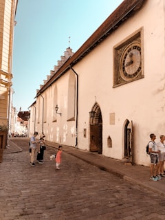 A historical street scene features a group of people, including a child in a red dress, standing on a cobblestone path. The path runs alongside a large beige medieval building with a prominent, intricate clock embedded in the wall. The building has architectural details typical of old European structures, with arched doorways and overhanging eaves. The sky is clear and blue, suggesting pleasant weather.