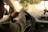 A person is driving a car, focusing on the road. The interior of the car is visible, including the steering wheel, dashboard, and radio display showing a station. The driver's arm is extended on the wheel, and there is a smartphone holder attached to the windshield.