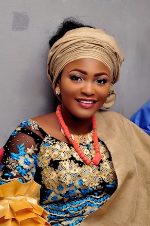 A woman wears traditional attire featuring intricate embroidery in gold, blue, and black. She accessorizes with a head wrap and coral beaded necklace, and holds a golden fabric or object.