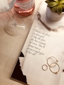 A wine glass and a pink wine bottle sit on a light wooden surface next to an open magazine displaying a quote. Beside the magazine, a small potted succulent plant is visible. Three delicate gold rings are placed on the magazine page.