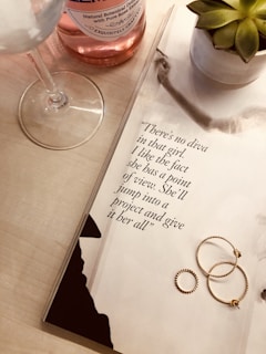 A wine glass and a pink wine bottle sit on a light wooden surface next to an open magazine displaying a quote. Beside the magazine, a small potted succulent plant is visible. Three delicate gold rings are placed on the magazine page.