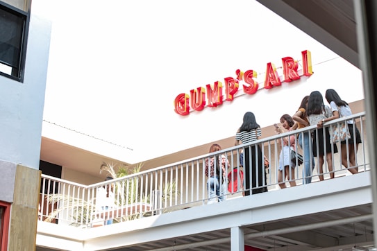 A group of young people is standing on a balcony, engaged in conversation and looking at their phones. They are near a large sign that reads 'GUMP'S ARI.' The setting appears to be an outdoor area with modern architecture. The scene is brightly lit, suggesting daytime.