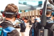 Two individuals wearing headphones with microphones are seen from behind. They appear to be in a motorsport setting, as indicated by the 'Racing Electronics' branding on their headsets. People in the background are blurred, suggesting a busy environment, likely at a racing event.