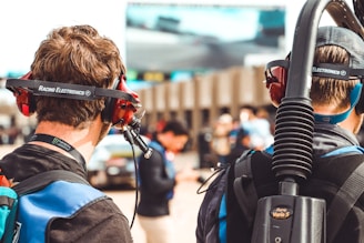Two individuals wearing headphones with microphones are seen from behind. They appear to be in a motorsport setting, as indicated by the 'Racing Electronics' branding on their headsets. People in the background are blurred, suggesting a busy environment, likely at a racing event.