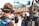 Two individuals wearing headphones with microphones are seen from behind. They appear to be in a motorsport setting, as indicated by the 'Racing Electronics' branding on their headsets. People in the background are blurred, suggesting a busy environment, likely at a racing event.