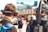 Two individuals wearing headphones with microphones are seen from behind. They appear to be in a motorsport setting, as indicated by the 'Racing Electronics' branding on their headsets. People in the background are blurred, suggesting a busy environment, likely at a racing event.
