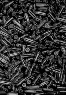 A large collection of metallic bullets tightly packed together. The bullets are shiny and appear to be of a similar size and shape, indicating they are likely of the same caliber. The image has high contrast, enhancing the reflective surfaces of the metal casings.