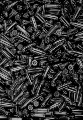 A large collection of metallic bullets tightly packed together. The bullets are shiny and appear to be of a similar size and shape, indicating they are likely of the same caliber. The image has high contrast, enhancing the reflective surfaces of the metal casings.