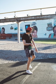 A person in casual attire, including a black t-shirt, red cap, patterned shorts, and white sneakers, poses playfully on a paved surface. The background features a large mural on a white building wall with colorful artwork depicting faces and abstract designs. There are large containers and a structure resembling an unfinished bridge or overpass.