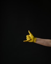A human hand painted yellow is making a gesture commonly known as the 'shaka' or 'hang loose' sign. The gesture is created by extending the thumb and little finger while the other fingers are curled. The background is solid black, providing a stark contrast to the brightly colored hand.