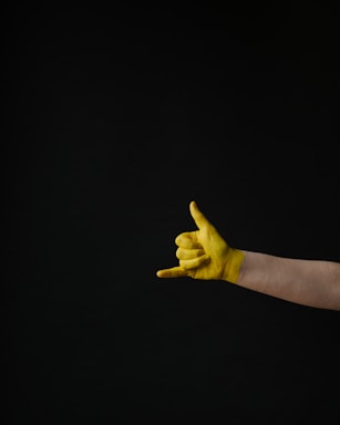 A human hand painted yellow is making a gesture commonly known as the 'shaka' or 'hang loose' sign. The gesture is created by extending the thumb and little finger while the other fingers are curled. The background is solid black, providing a stark contrast to the brightly colored hand.