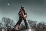 A large sculpture of two hands pressed together in a praying position stands prominently against a backdrop of leafless trees and a night sky featuring a visible moon.