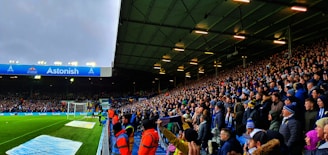 A crowded stadium filled with enthusiastic spectators. Many are standing and appear to be watching a sporting event with intensity. The stadium lighting is on, and the field is partially visible to the left. Some people are wearing team apparel, suggesting a sports team atmosphere. There are stewards wearing bright orange jackets along the edge of the field.