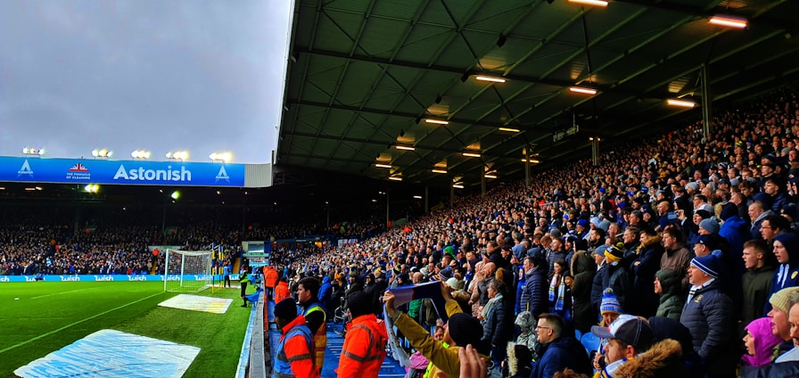 A crowded stadium filled with enthusiastic spectators. Many are standing and appear to be watching a sporting event with intensity. The stadium lighting is on, and the field is partially visible to the left. Some people are wearing team apparel, suggesting a sports team atmosphere. There are stewards wearing bright orange jackets along the edge of the field.