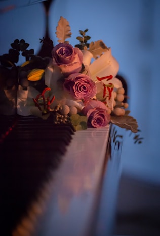 A bouquet of flowers featuring prominently purple roses and white lilies, surrounded by a variety of leaves and smaller blooms, rests elegantly on a highly polished surface that resembles a piano. The scene is set in dim lighting, creating a serene and intimate atmosphere.