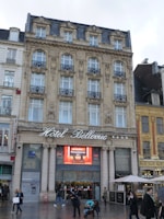 A historic multi-story building with ornate stonework and decorative iron railings on the windows. The words 'Hôtel Bellevue' are prominently displayed above the entrance. People are walking in front of the building, one person pushing a stroller. The architecture is classic with intricate details around the windows and a mansard roof.