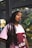 A person with long braided hair leaning against a dark vertical pole in what appears to be an outdoor setting. They are wearing a pink and maroon Adidas shirt with text on it. There is a colorful, abstract mural in the blurred background.