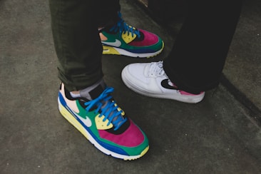 Two pairs of sneakers with contrasting designs are placed on a concrete surface. One pair features a colorful combination of pink, green, blue, and yellow, while the other pair is mainly white with pink and black accents. The different styles and colors create a vibrant visual contrast.