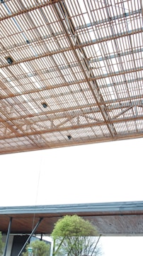 A large, modern architectural structure features a ceiling made of interwoven wooden slats, creating an intricate lattice pattern. Below, an overhang with a flat wooden underside is supported by metal beams. In the lower part of the image, green trees are visible, adding a touch of nature to the scene.