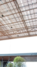 A large, modern architectural structure features a ceiling made of interwoven wooden slats, creating an intricate lattice pattern. Below, an overhang with a flat wooden underside is supported by metal beams. In the lower part of the image, green trees are visible, adding a touch of nature to the scene.