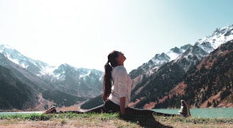 A person is performing a yoga split on a grassy area near a serene lake, surrounded by snow-capped mountains under a clear sky. The individual is dressed in a white top and dark pants, with long hair tied back. The scene exudes tranquility and connection with nature.