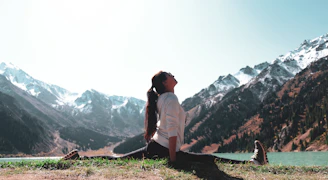 A person is performing a yoga split on a grassy area near a serene lake, surrounded by snow-capped mountains under a clear sky. The individual is dressed in a white top and dark pants, with long hair tied back. The scene exudes tranquility and connection with nature.