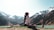 A person is performing a yoga split on a grassy area near a serene lake, surrounded by snow-capped mountains under a clear sky. The individual is dressed in a white top and dark pants, with long hair tied back. The scene exudes tranquility and connection with nature.