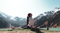 A person is performing a yoga split on a grassy area near a serene lake, surrounded by snow-capped mountains under a clear sky. The individual is dressed in a white top and dark pants, with long hair tied back. The scene exudes tranquility and connection with nature.
