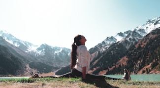 A person is performing a yoga split on a grassy area near a serene lake, surrounded by snow-capped mountains under a clear sky. The individual is dressed in a white top and dark pants, with long hair tied back. The scene exudes tranquility and connection with nature.