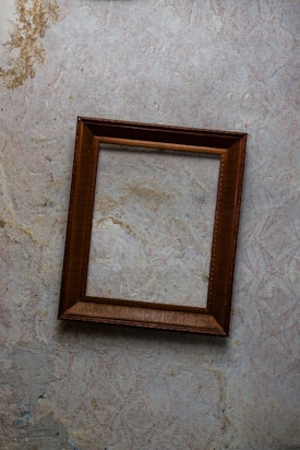 An empty, weathered wooden picture frame hangs slightly askew on an aged and peeling, patterned wall. The wall shows signs of significant wear and tear with parts of the design faded or flaking off.