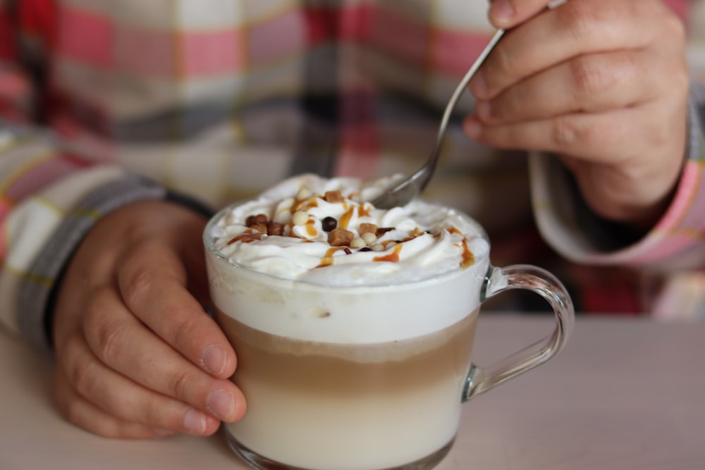 person holding ice cream with white cream on clear glass mug