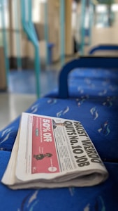 A folded newspaper lies on a blue patterned train seat. The newspaper's headline is partially visible, reading 'QUITS No. 10 JOB.' The background reveals a train interior, with blue and turquoise accents.