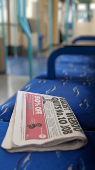 A folded newspaper lies on a blue patterned train seat. The newspaper's headline is partially visible, reading 'QUITS No. 10 JOB.' The background reveals a train interior, with blue and turquoise accents.