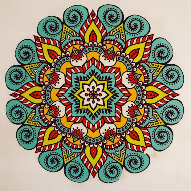 A vibrant, colorful mandala featuring intricate patterns and symmetrical designs. The mandala has a central flower-like motif surrounded by layers of geometric and floral shapes. The design is characterized by the use of bold lines and a variety of colors, creating a visually striking appearance.