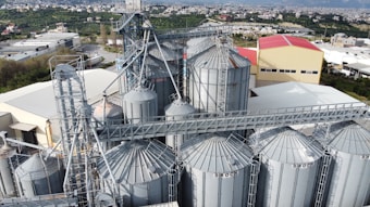 An industrial complex featuring multiple large grain silos with metal frameworks and a network of connecting conveyor systems. A backdrop of green fields and a town with mountainous terrain is visible.