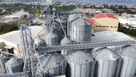 An industrial complex featuring multiple large grain silos with metal frameworks and a network of connecting conveyor systems. A backdrop of green fields and a town with mountainous terrain is visible.