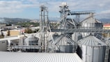A complex industrial facility with multiple large metal silos and various interconnected pipes and structures, set against a backdrop of distant hills and a clear sky. The surrounding area includes other industrial buildings and green landscapes.